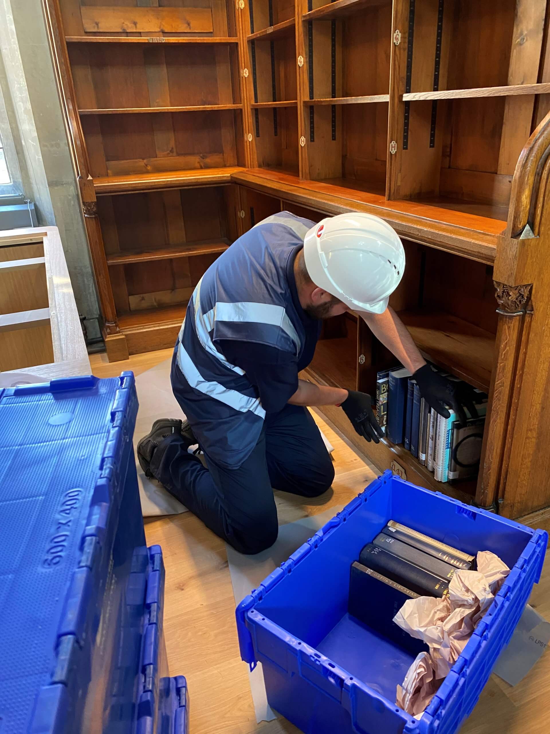 Person wearing hardhat and construction vest putting books from crate into a largely empty bookshelf